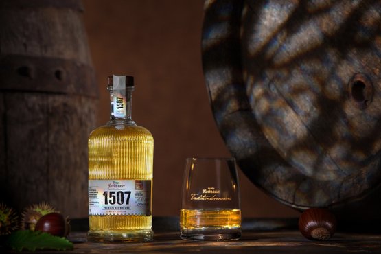 Flasche „1507“ mit goldenem Destillat neben Glas auf Holztisch, im Hintergrund Holzfässer.