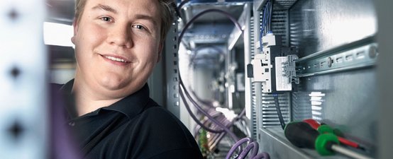 In einem Schaltschrank lächelt ein junger Mann, während er mit Werkzeugen und Kabeln arbeitet, die ordentlich angeordnet sind.