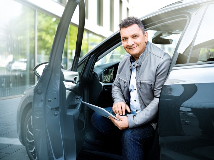 With a smile on his face, the man sits in his car and types on his laptop, surrounded by modern buildings.