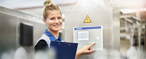 In einer hellen Fabrik lächelt eine junge Frau, während sie eine Maschine mit einem Steuergerät bedient.