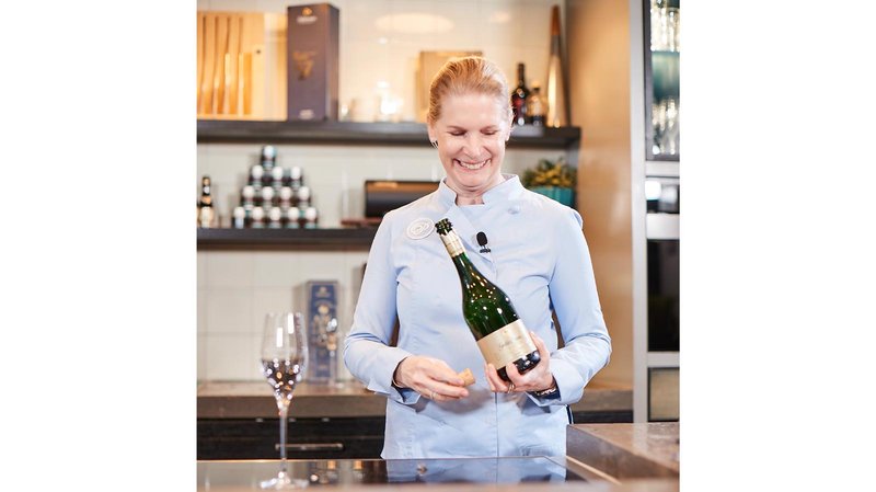 Eine lächelnde Frau in einer blauen Bluse hält eine Sektflasche und einen Korken in einer modernen Küche.