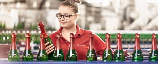 Eine Frau mit Brille betrachtet eine Flasche Sekt in einer Produktionsstätte, umgeben von vielen weiteren Flaschen.