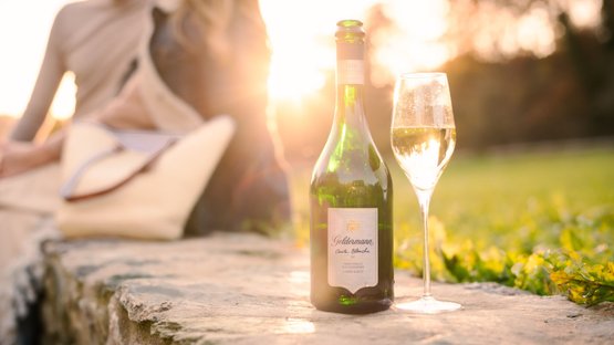 Geldermann: Eine grüne Sektflasche steht auf einem Stein, daneben ein Glas mit sprudelndem Sekt, während die Sonne untergeht.
