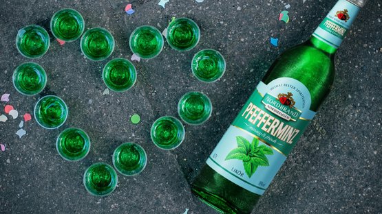 Pfeffi: Die grüne Flasche Pfefferminzlikör steht neben mehreren Schnapsgläsern, während farbenfrohes Konfetti den Boden schmückt.
