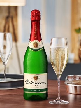 Eine Flasche Rotkäppchen Sekt steht auf einem Tisch, daneben ein Glas mit sprudelndem Sekt und weiteren Gläsern im Hintergrund.