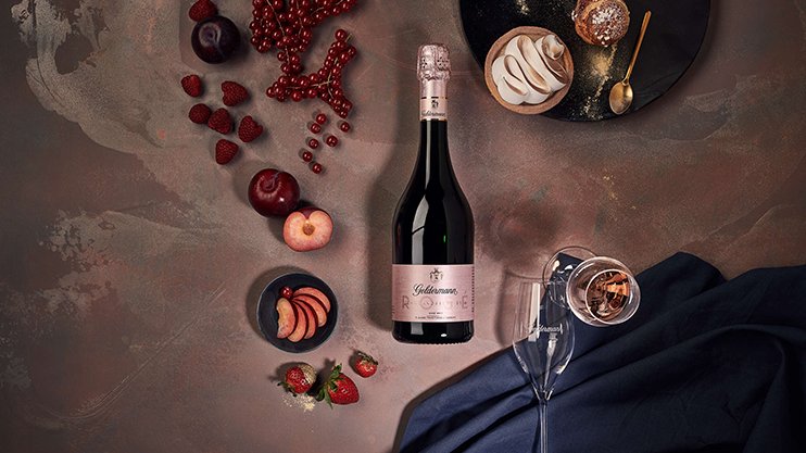 Ein eleganter Tisch mit einer Flasche Rosé-Sekt, frischen Früchten und einem Dessert auf einem dunklen Hintergrund.