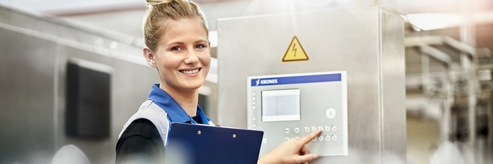 Eine lächelnde Frau in Arbeitskleidung bedient ein Steuergerät in einer modernen Industrieumgebung.