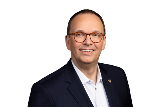 Frank Albers: A smiling man with glasses wears a dark suit and a white shirt, against a neutral background.