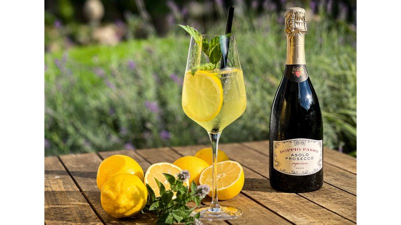 Ein Glas mit sprudelndem Getränk, garniert mit Zitronen und Minze, neben einer Flasche Prosecco auf einem rustikalen Tisch.