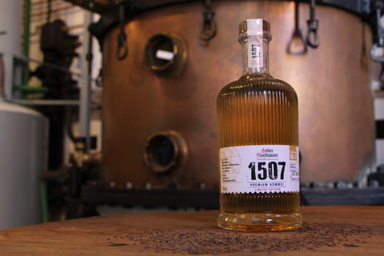 Eine Flasche Echter Nordhäuser Kümmel auf einem dunklen Holztisch vor einer Brennblase aus Kupfer