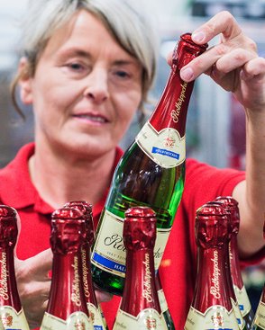 A woman in a red shirt lifts a bottle of sparkling wine with a red cork from a row of bottles on a table.