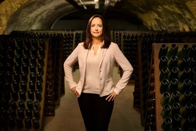 A confident woman stands in a wine cellar, surrounded by shelves full of wine bottles, with a determined look.