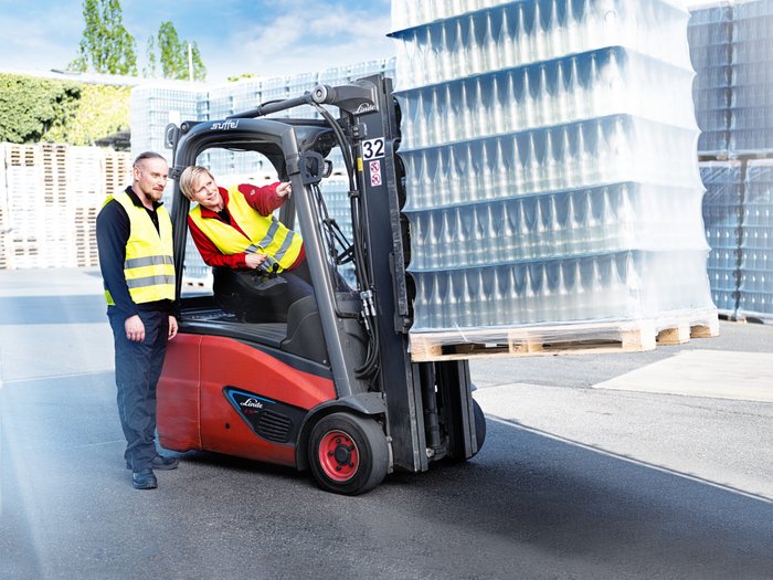 A woman in a forklift points at something while a man in safety clothing watches with interest, surrounded by bottles.