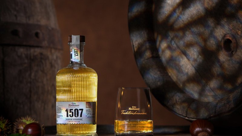 Flasche „1507“ mit goldenem Destillat neben Glas auf Holztisch, im Hintergrund Holzfässer.