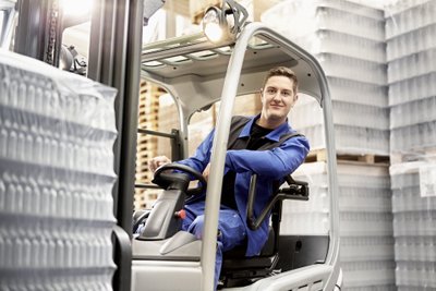 A young man in blue work clothes sits smiling in a forklift, surrounded by pallets and storage shelves.