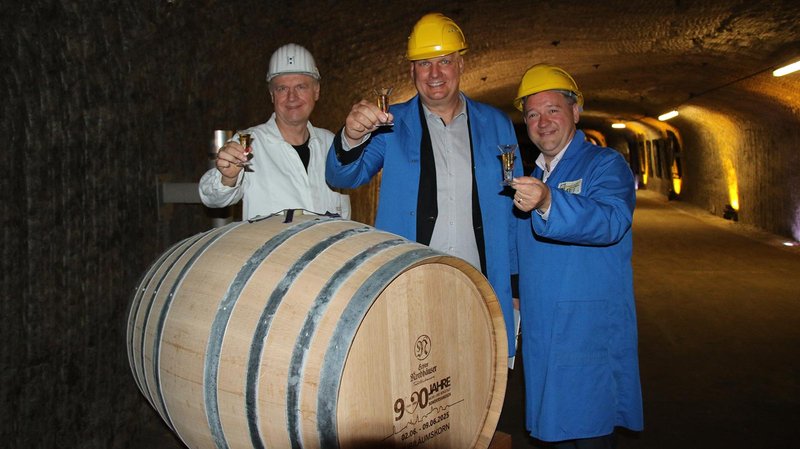 Drei Männer in Schutzkleidung und Helmen heben Gläser in einer unterirdischen Lagerhalle mit einem großen Holzfass.