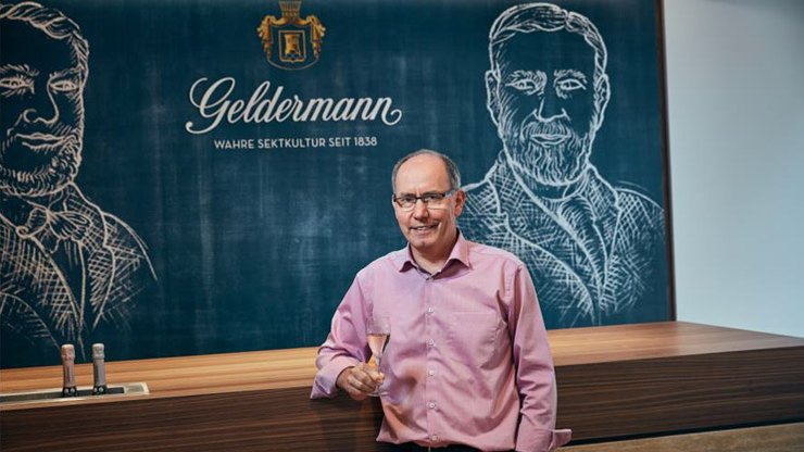 Der Mann mit Brille und rosa Hemd präsentiert stolz ein Glas Sekt vor einer Wand mit dem Schriftzug 'Geldermann'.
