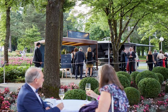 Elegante Gäste unterhalten sich in einem Park, während sie an einem Getränkestand stehen, umgeben von bunten Blumenbeeten.