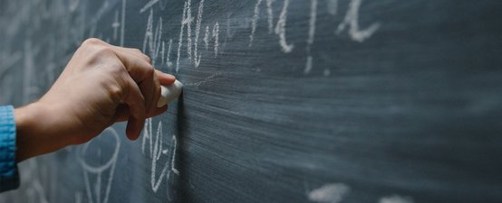 Eine Hand hält ein Stück Kreide und schreibt auf eine dunkle Tafel, während mathematische Symbole sichtbar sind.