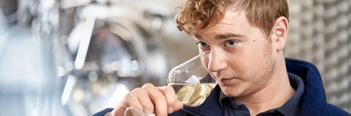 Der Mann mit dem lockigen Haar genießt einen Moment der Ruhe, während er Wein aus einem Glas in einer modernen Weinkellerei probiert.