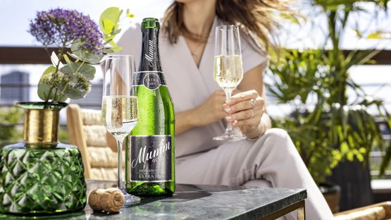 A woman wearing sunglasses enjoys Mumm sparkling wine on a sunny terrace, surrounded by plants and an elegant table.