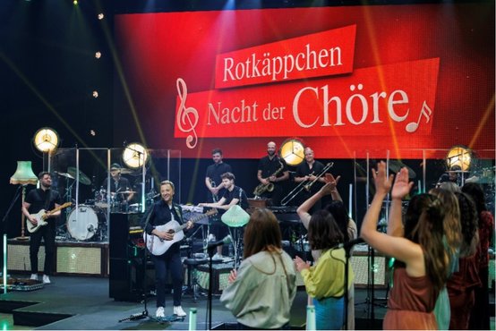 Ein großes Banner mit 'Nacht der Chöre' im Hintergrund, während die Musiker und das Publikum eine energiegeladene Atmosphäre genießen.