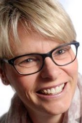 Eine lächelnde Frau mit blonden, kurzen Haaren und Brille, die in einem hellen Raum posiert.