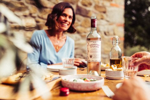 Eine fröhliche Frau sitzt an einem Tisch im Freien, umgeben von köstlichem Essen und einer Flasche Doppio Passo Wein.
