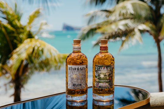 Zwei Flaschen Star & Key Rum auf einem Tisch vor tropischem Strand mit Palmen und türkisfarbenem Meer im Hintergrund.