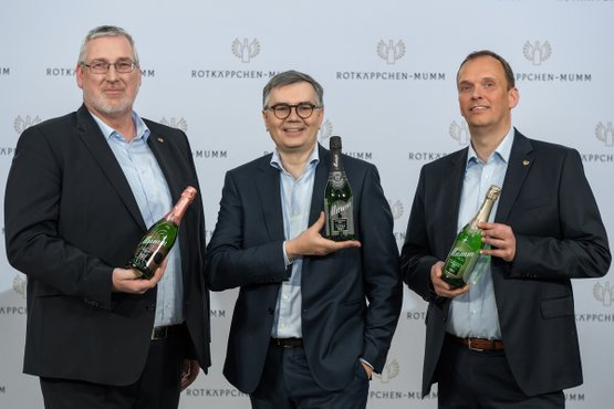 The three men, dressed in formal suits, smile and hold their champagne bottles in front of a striking background wall.