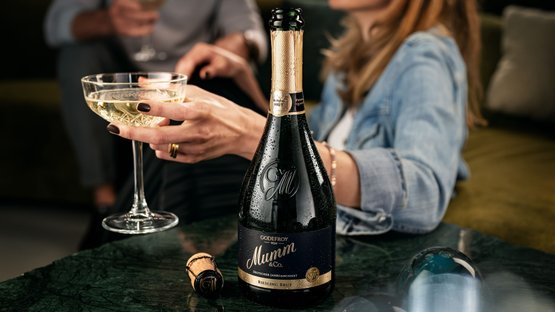 A woman holds a glass of sparkling wine in her hand while a bottle of Mumm Riesling Brut sits on the table.