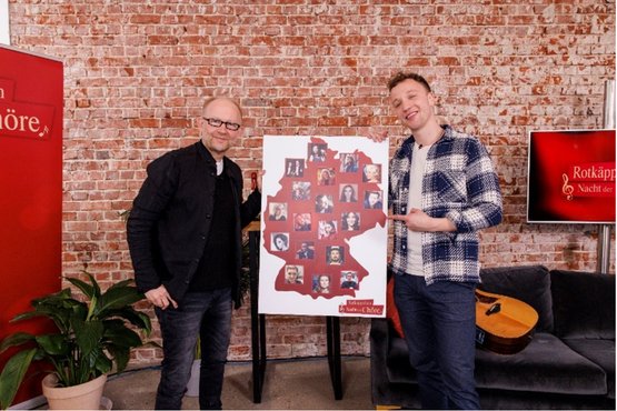 Zwei Männer stehen vor einer roten Backsteinwand und präsentieren ein Plakat mit Bildern von berühmten Persönlichkeiten in Deutschland.