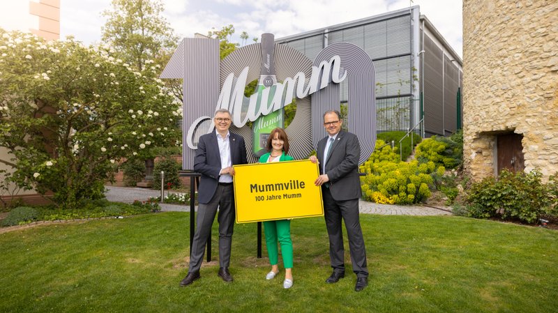 Drei Personen stehen im Freien vor einem großen Mumm-Schild, das 100 Jahre Mumm feiert, und halten ein gelbes Schild mit 'Mummville'.