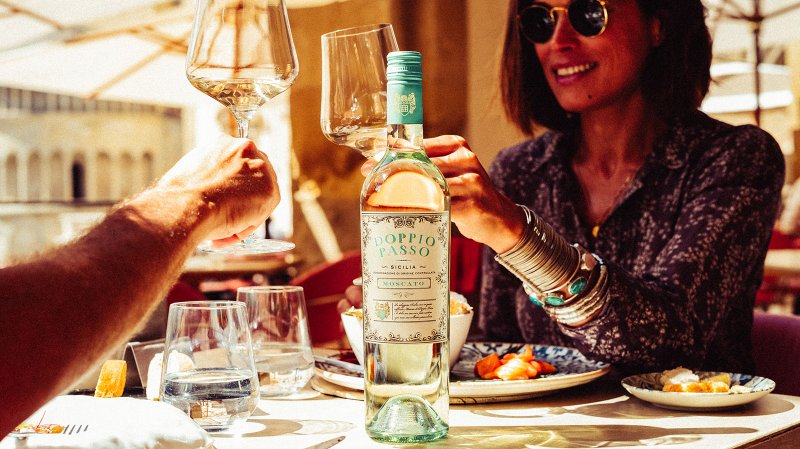 Ein Mann und eine Frau stoßen mit Weingläsern an, während eine Flasche Doppio Passo Moscato auf dem Tisch steht.