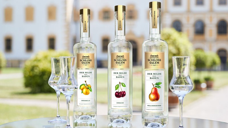 Drei Flaschen Schloss Salem Obstbrand stehen auf einem Tisch, umgeben von einer grünen Gartenlandschaft und Gläsern.