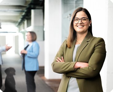A confident woman in an olive green blazer smiles while interacting with other people in a modern office.
