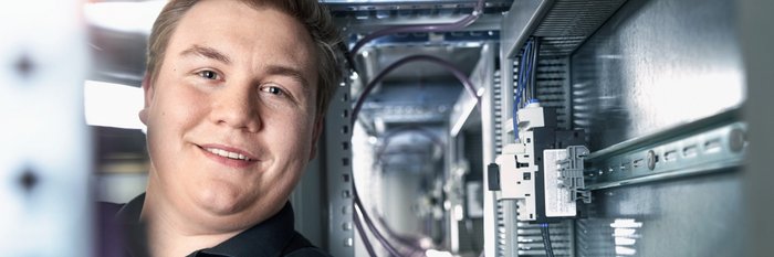 In einem modernen Technikraum lächelt ein junger Mann, während er an einem Schaltschrank mit verschiedenen Kabeln und Geräten arbeitet.