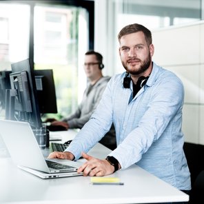 Der Mann im Vordergrund trägt ein hellblaues Hemd und schaut direkt in die Kamera, während er an seinem Laptop arbeitet.