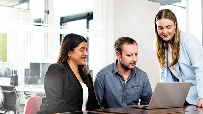 Three people are sitting at a table, looking at a laptop together while having an animated conversation.
