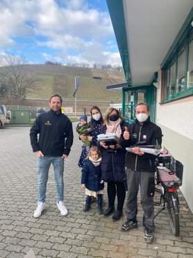Eine Gruppe von fünf Personen steht vor einem Gebäude, alle tragen Masken und lächeln, während sie verschiedene Gegenstände halten.