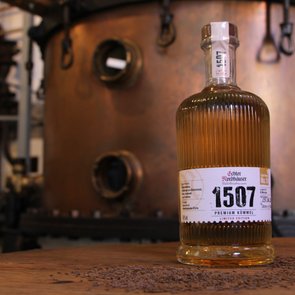 Eine Flasche Echter Nordhäuser Kümmel auf einem dunklen Holztisch vor einer Brennblase aus Kupfer