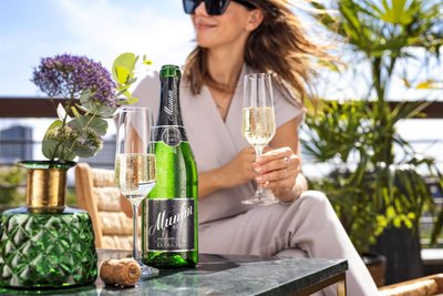 A woman wearing sunglasses enjoys Mumm sparkling wine on a sunny terrace, surrounded by plants and an elegant table.