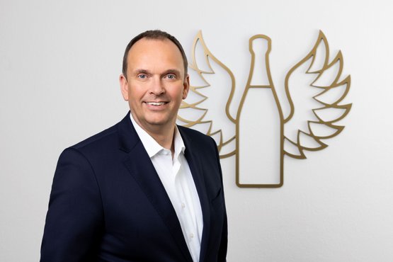 Frank Albers: A smiling man in a dark suit stands in front of a wall with the golden wing logo of Rotkäppchen Mumm.