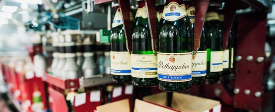 Eine Reihe von Flaschen Rotkäppchen Sekt hängt in einer Produktionslinie, umgeben von verschwommenen Maschinen und Lichtern.