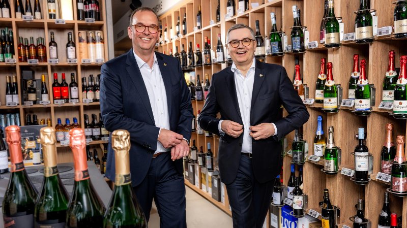 Zwei Männer in Anzügen lächeln vor einem Regal voller Weinflaschen und Sektflaschen in einem Geschäft.