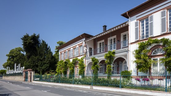 Eltville: An elegant building with a balcony, surrounded by lush greenery and colorful flowers, under a bright blue sky.