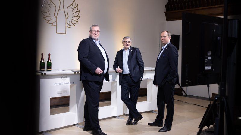 Drei Männer in Anzügen posieren lässig vor einem eleganten Hintergrund mit einem Logo und Flaschen Sekt auf einem Tisch.