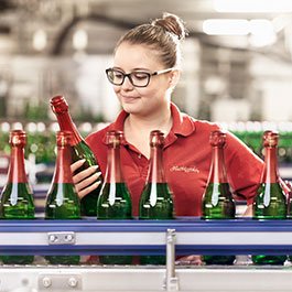 Eine Frau mit Brille inspiziert eine Sektflasche in einer Produktionslinie, umgeben von weiteren Flaschen in grünen Glasbehältern.