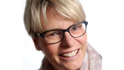 A smiling woman with short blonde hair and glasses posing in a bright room.