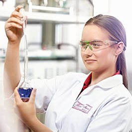 Eine junge Frau in einem Labor hält eine Pipette und betrachtet eine blaue Flüssigkeit in einem Erlenmeyerkolben.